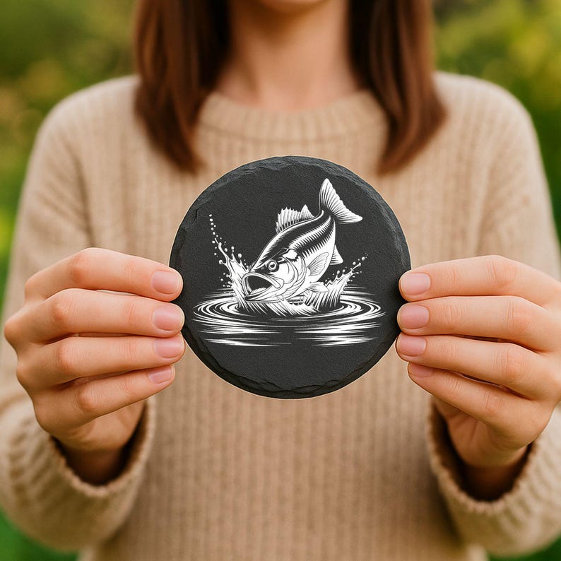 May include: A round, black slate coaster featuring a white graphic of a fish leaping out of water. The fish is detailed with scales and fins, and the water is depicted with concentric circles and splashes. The coaster is held by a person.