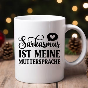 May include: White ceramic mug with a black handle. The mug features the text "Sarkasmus IST MEINE MUTTERSPRACHE" in black, with a heart symbol. The mug is likely for coffee or tea.