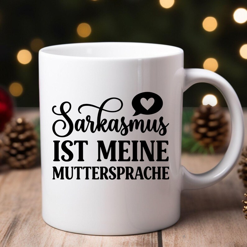 May include: White ceramic mug with a black handle. The mug features the text "Sarkasmus IST MEINE MUTTERSPRACHE" in black, with a heart symbol. The mug is likely for coffee or tea.