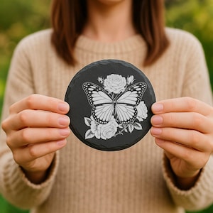 Puede incluir: Un posavasos redondo de pizarra negra con un dise&ntilde;o de mariposa blanca y rosas blancas. El posavasos est&aacute; sostenido por una persona, destacando el grabado detallado y los colores contrastantes. Ideal para la decoraci&oacute;n del hogar.