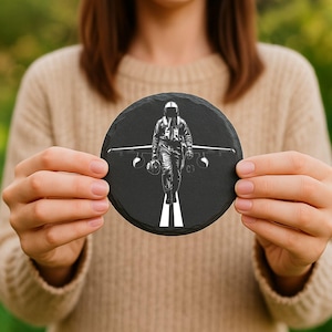 May include: A round, black slate coaster featuring a white graphic of a pilot walking towards the viewer, with an airplane behind him. The pilot is wearing a flight suit and helmet. The coaster is held in front of a blurred background.