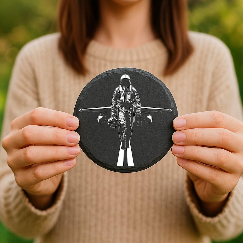 May include: A round, black slate coaster featuring a white graphic of a pilot walking towards the viewer, with an airplane behind him. The pilot is wearing a flight suit and helmet. The coaster is held in front of a blurred background.