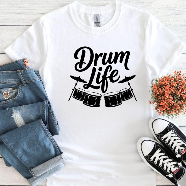 May include: White t-shirt with the words "Drum Life" in a black, stylized font, along with a drum set graphic. The shirt is paired with blue jeans and black and white sneakers.