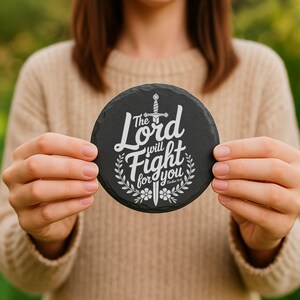 Puede incluir: Un posavasos redondo de pizarra negra con texto blanco que dice "The Lord will Fight for you". El dise&ntilde;o incluye una espada, elementos florales y el vers&iacute;culo b&iacute;blico &Eacute;xodo 14:14. El posavasos se sostiene frente a un fondo borroso.