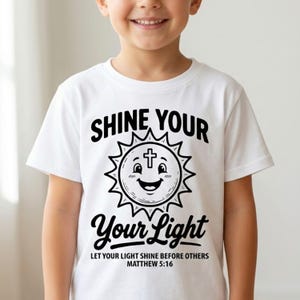 May include: White t-shirt with the text "SHINE YOUR LIGHT" in black, featuring a smiling sun with a cross. The shirt also includes the text "LET YOUR LIGHT SHINE BEFORE OTHERS MATTHEW 5:16".