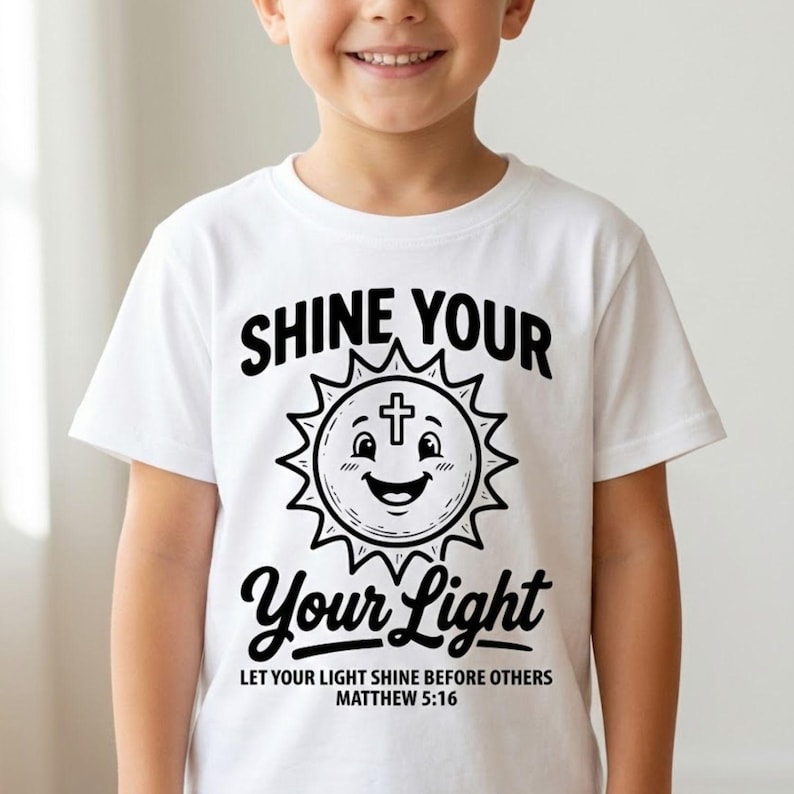 May include: White t-shirt with the text "SHINE YOUR LIGHT" in black, featuring a smiling sun with a cross. The shirt also includes the text "LET YOUR LIGHT SHINE BEFORE OTHERS MATTHEW 5:16".