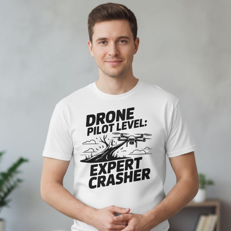 May include: White t-shirt with a black graphic of a drone and the text "DRONE PILOT LEVEL: EXPERT CRASHER". The design includes a drone, a road, and clouds. The shirt is worn by a person.