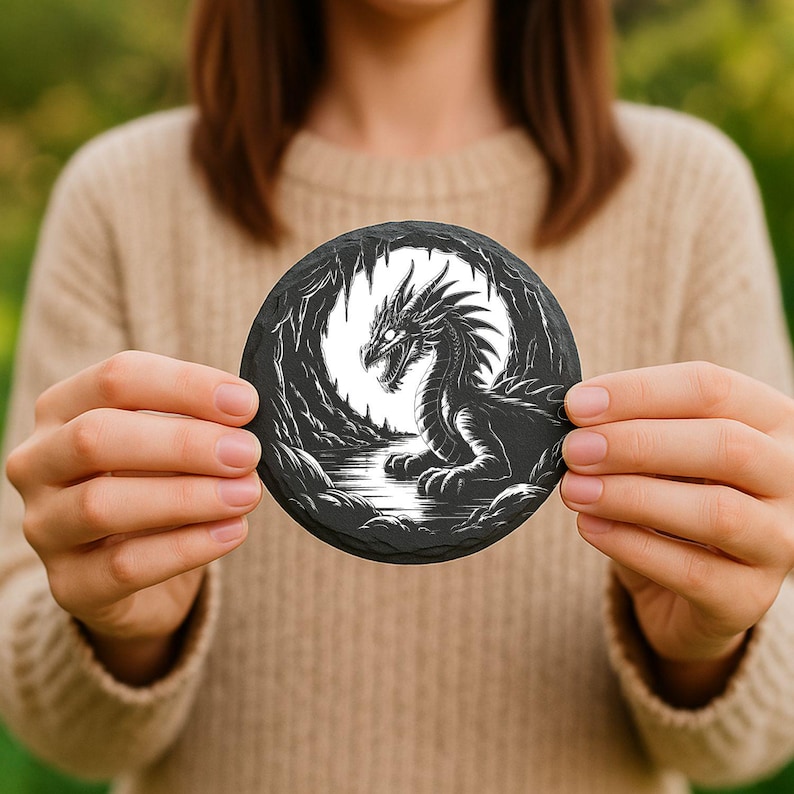 May include: A round, black coaster featuring a detailed illustration of a dragon in a cave setting. The dragon is black and white, with intricate scales and a fierce expression. The coaster is held in front of a blurred green background.