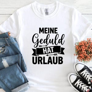 May include: White crew neck t-shirt with the German phrase "MEINE Geduld HAT URLAUB" in black script. The shirt is paired with blue jeans and black and white sneakers.