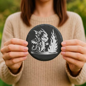 May include: A round, black slate coaster featuring a white illustration of a cartoon dragon and a flame. The dragon has wings and is standing next to the flame. The coaster is held in front of a person wearing a beige sweater.