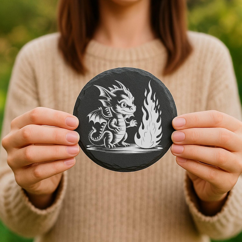 May include: A round, black slate coaster featuring a white illustration of a cartoon dragon and a flame. The dragon has wings and is standing next to the flame. The coaster is held in front of a person wearing a beige sweater.