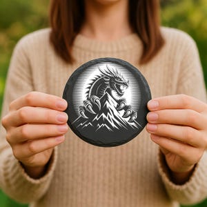 May include: A round, black slate coaster featuring a detailed white illustration of a dragon perched atop a mountain range. The dragon has sharp claws and a menacing expression. The coaster is held in front of a blurred background.