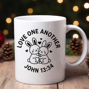 May include: White ceramic mug featuring a black graphic of two hugging bunnies with hearts. The text "LOVE ONE ANOTHER" and "JOHN 13:34" are also in black. The mug has a curved handle and is set against a blurred background.