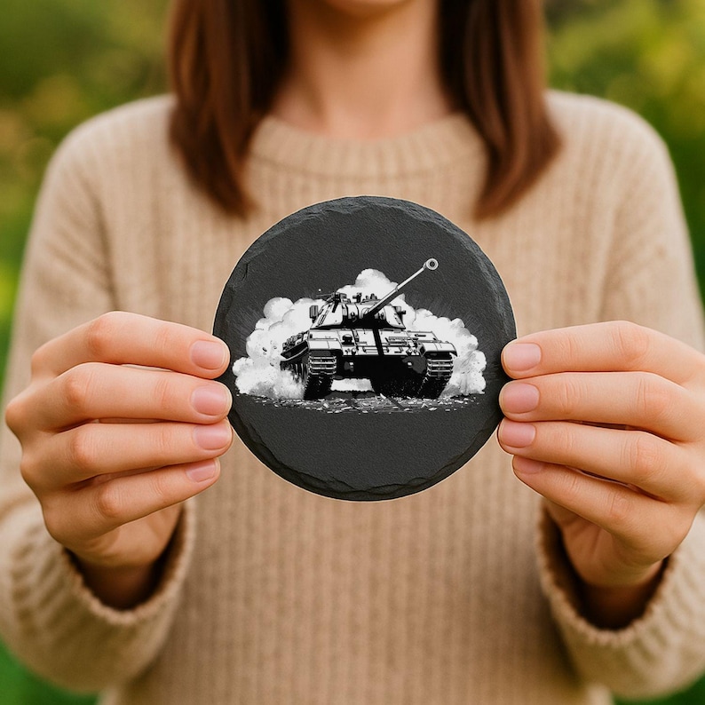 May include: A round, black slate coaster featuring a detailed black and white illustration of a tank emerging from a cloud of smoke. The coaster is held up by a person.