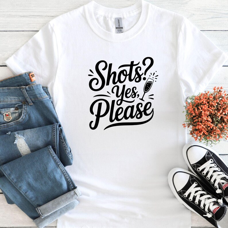 May include: White t-shirt with black text that reads "Shots? Yes, Please" and a black silhouette of a champagne flute.