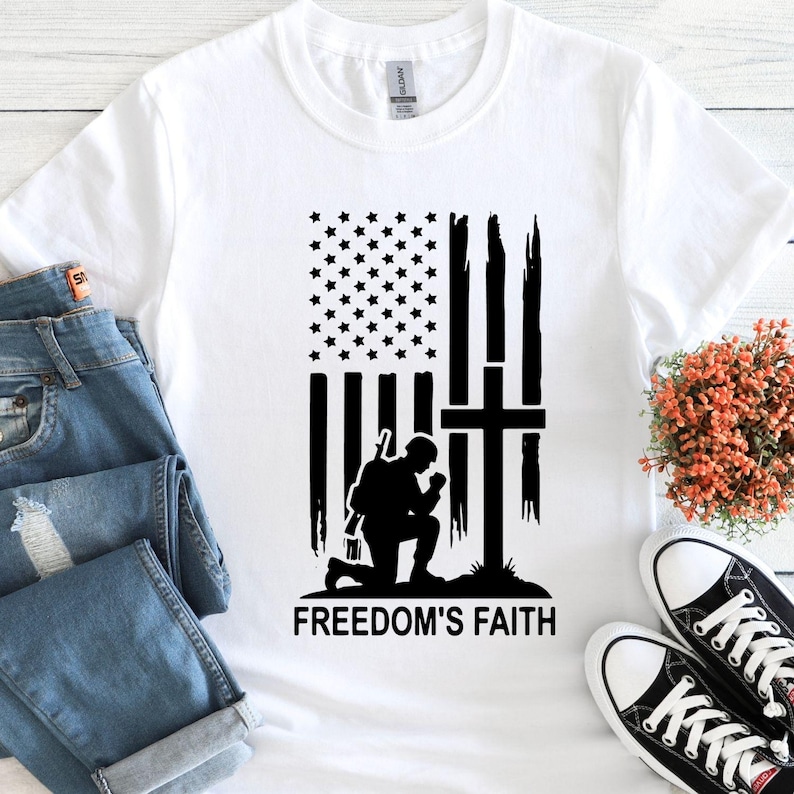 May include: White t-shirt featuring a black graphic of an American flag with a cross and a kneeling soldier. The text "FREEDOM'S FAITH" is printed below the graphic. The shirt is paired with blue jeans and black canvas sneakers.