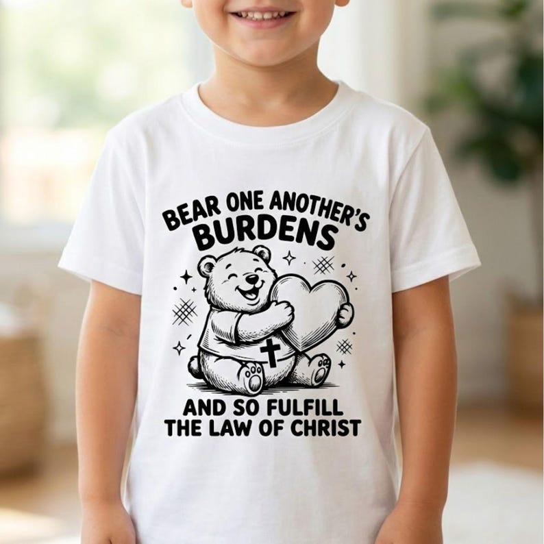 May include: White t-shirt featuring a smiling bear holding a heart. The bear has a cross on its shirt. The text on the shirt reads "Bear one another's burdens and so fulfill the law of Christ."