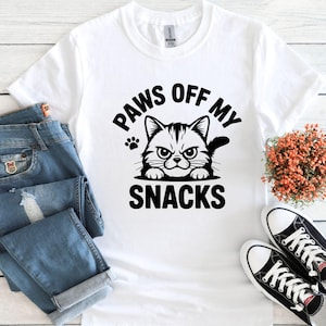 May include: White t-shirt with the text "PAWS OFF MY SNACKS" and a graphic of a grumpy cat. The shirt is paired with blue jeans and black and white sneakers, suggesting a casual style.