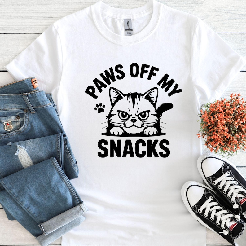 May include: White t-shirt with the text "PAWS OFF MY SNACKS" and a graphic of a grumpy cat. The shirt is paired with blue jeans and black and white sneakers, suggesting a casual style.