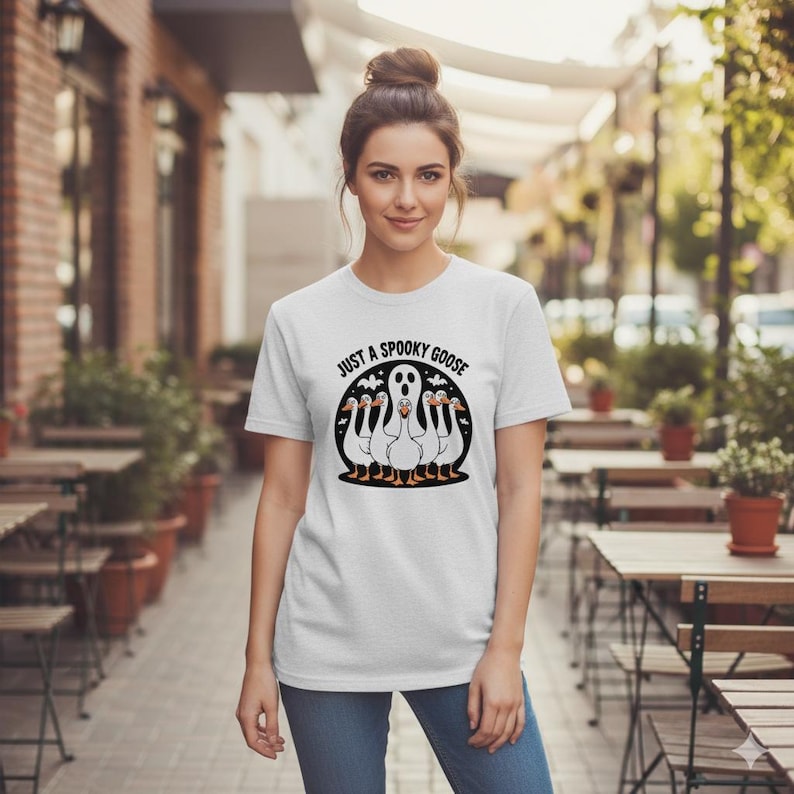 May include: A light grey t-shirt with the text "JUST A SPOOKY GOOSE" in a black circle. The graphic features a ghost and five geese. The geese have orange beaks and feet. The shirt is made of a soft material.