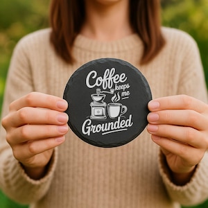 May include: A round, black slate coaster with the words "Coffee keeps me Grounded" in white script. The coaster features a coffee grinder and a coffee cup graphic. The coaster is held in front of a blurred background.
