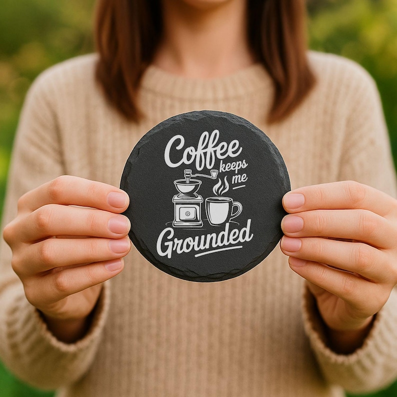 May include: A round, black slate coaster with the words "Coffee keeps me Grounded" in white script. The coaster features a coffee grinder and a coffee cup graphic. The coaster is held in front of a blurred background.