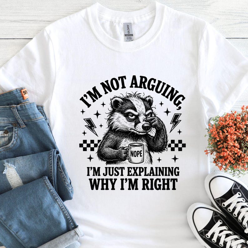K&ouml;nnte beinhalten: Wei&szlig;es T-Shirt mit einem schwarz-wei&szlig;en Dachs, der eine Tasse mit der Aufschrift "NOPE" h&auml;lt. Der Text lautet: "ICH STREITE NICHT, ICH ERKL&Auml;RE NUR, WARUM ICH RECHT HABE". Das Design beinhaltet Blitze und Sterne.
