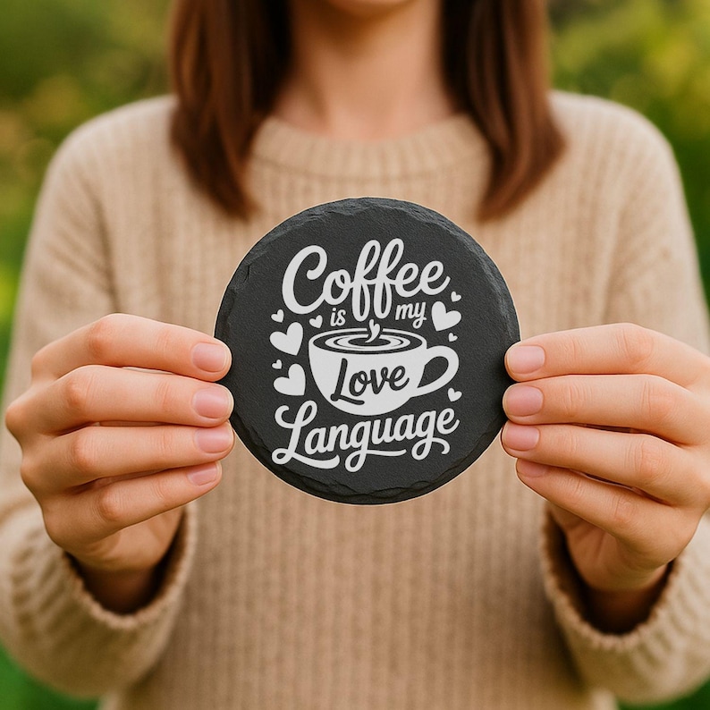 May include: A round, black slate coaster with the phrase "Coffee is my Love Language" in white script and a coffee cup graphic. The coaster is held up by a person.