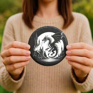 May include: Round black slate coaster featuring a white illustration of a dragon breathing fire in front of a castle and a full moon. The coaster is held by a person.