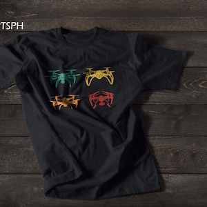 Könnte beinhalten: Schwarzes T-Shirt mit vier Retro-Drohnen-Designs in verschiedenen Farben. Die Drohnen sind in einem Rastermuster auf der Vorderseite des Shirts angeordnet.