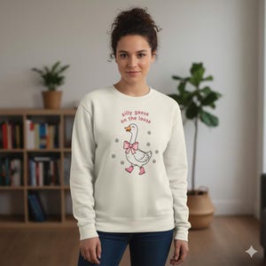 May include: Cream-coloured sweatshirt with the text "silly goose on the loose" in pink. The graphic features a cartoon goose wearing a pink bow, pink boots, and small flowers. The sweatshirt is a casual, comfortable style.