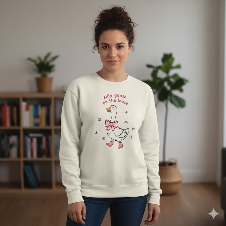 May include: Cream-coloured sweatshirt with the text "silly goose on the loose" in pink. The graphic features a cartoon goose wearing a pink bow, pink boots, and small flowers. The sweatshirt is a casual, comfortable style.
