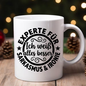 May include: White ceramic mug with a black circular design that reads "EXPERTE F&Uuml;R SARKASMUS & IRONIE" and "Ich wei&szlig; alles besser!" in a decorative font. The mug has a curved handle and is set against a blurred background.
