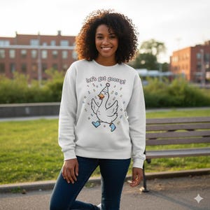May include: A light grey sweatshirt featuring the text "Let's get goosey!" and a cartoon goose wearing a party hat and blue boots. The goose is surrounded by confetti. The person is wearing blue jeans.