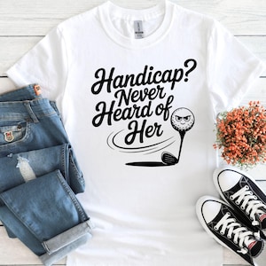May include: White t-shirt with the text "Handicap? Never Heard of Her" in black script. A golf ball with a face is on a tee, with a golf club below. The shirt is paired with blue jeans and black and white sneakers.
