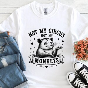 K&ouml;nnte beinhalten: Wei&szlig;es T-Shirt mit einer schwarzen Grafik eines Opossums und dem Text "NOT MY CIRCUS NOT MY MONKEYS". Das Design beinhaltet Sterne und Herzen. Das Shirt wird mit blauen Jeans und schwarz-wei&szlig;en Turnschuhen gezeigt.