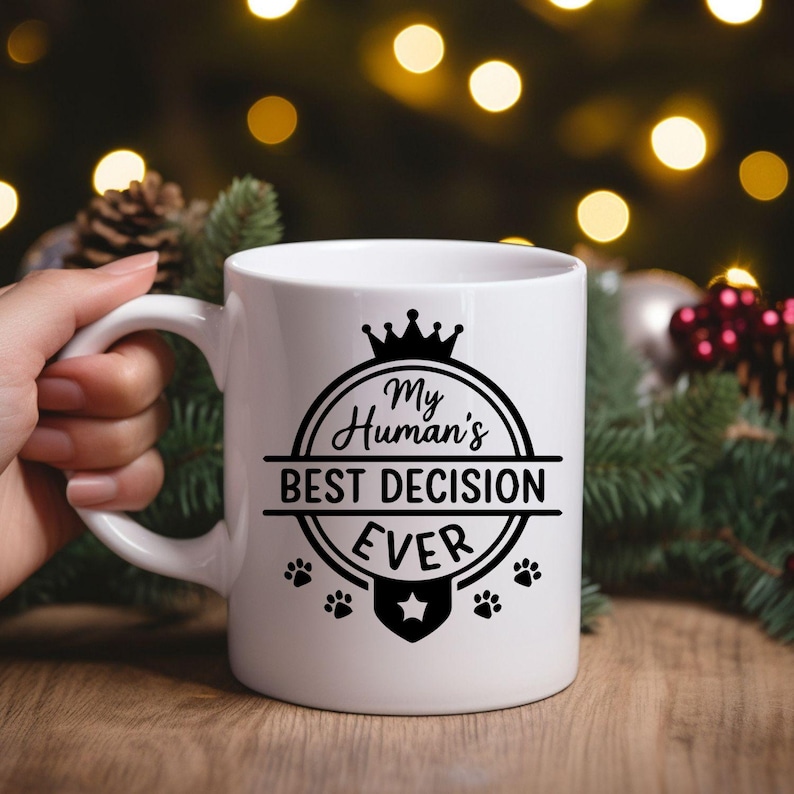 May include: White ceramic mug with a black graphic that reads "My Human's BEST DECISION EVER." The design includes a crown, paw prints, and a star. The mug is held by a hand, with a blurred background of Christmas decorations.