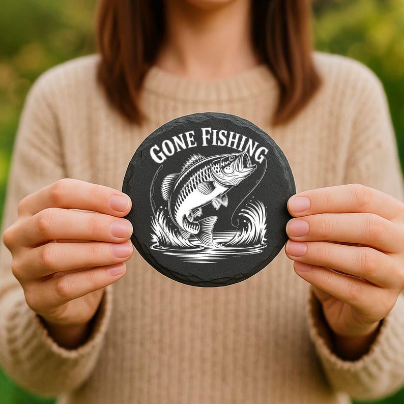 May include: Round black slate coaster with the words "GONE FISHING" in white, along with a detailed illustration of a fish jumping out of the water. The coaster is held in front of a blurred green background.