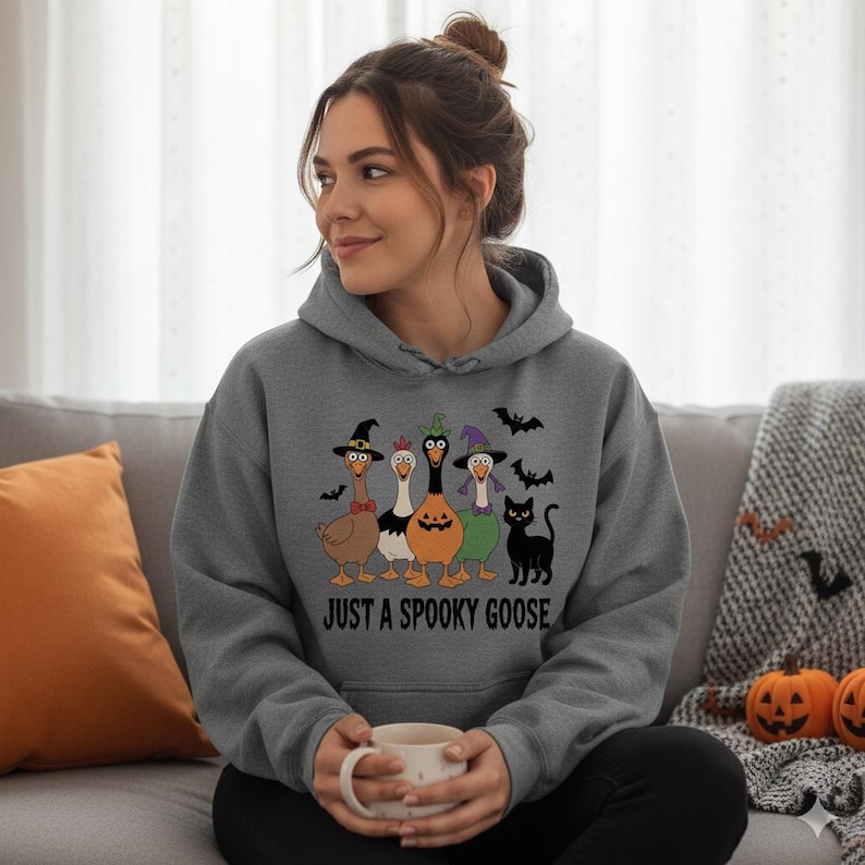 May include: A grey hoodie with a Halloween design. The graphic shows five cartoon geese wearing witch hats, a pumpkin, and a black cat, with bats overhead. The text "JUST A SPOOKY GOOSE" is printed below the geese, perfect for the spooky season.