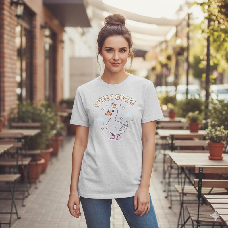 May include: A light grey t-shirt with a cartoon goose wearing a crown and pink shoes, and the text "QUEEN GOOSE" above. The design includes clouds and sparkles. The shirt is made of a soft material.