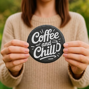 May include: A round, black slate coaster with the words "Coffee and Chill" in white script. The coaster is held by a person wearing a beige sweater. The design includes a coffee cup graphic.