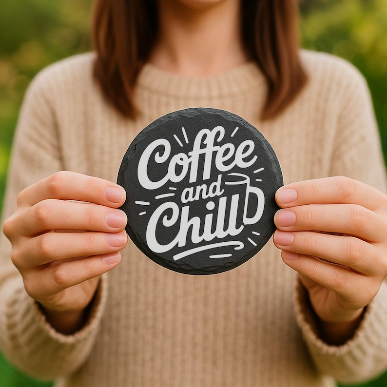 May include: A round, black slate coaster with the words "Coffee and Chill" in white script. The coaster is held by a person wearing a beige sweater. The design includes a coffee cup graphic.