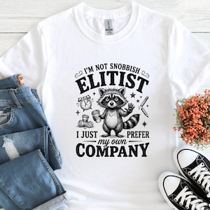 K&ouml;nnte beinhalten: Wei&szlig;es T-Shirt mit einem Waschb&auml;ren-Motiv, das eine Kaffeetasse h&auml;lt, und dem Text: &bdquo;Ich bin nicht snobistisch, elit&auml;r, ich bevorzuge einfach meine eigene Gesellschaft.&ldquo; Das Design umfasst eine Uhr, Sterne und einen Bleistift.