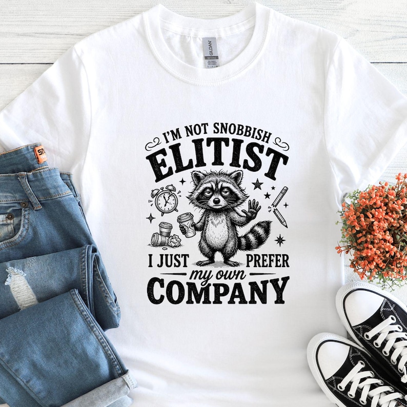 K&ouml;nnte beinhalten: Wei&szlig;es T-Shirt mit einem Waschb&auml;ren-Motiv, das eine Kaffeetasse h&auml;lt, und dem Text: &bdquo;Ich bin nicht snobistisch, elit&auml;r, ich bevorzuge einfach meine eigene Gesellschaft.&ldquo; Das Design umfasst eine Uhr, Sterne und einen Bleistift.
