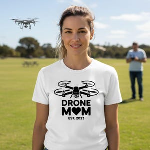 May include: White t-shirt with a black drone graphic and the words "DRONE MOM" with a heart and "EST. 2023". A drone is flying in the background. A person is standing in the background.