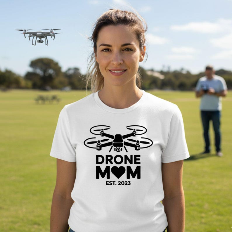 May include: White t-shirt with a black drone graphic and the words "DRONE MOM" with a heart and "EST. 2023". A drone is flying in the background. A person is standing in the background.