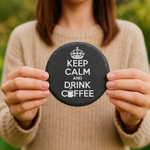 May include: A round, black slate coaster with the words "KEEP CALM AND DRINK COFFEE" in white, along with a crown and coffee cup graphic. The coaster is held up by a person.