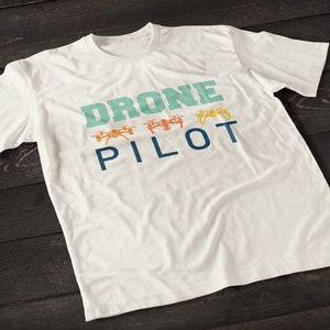 Puede incluir: Camiseta blanca con el texto "DRONE PILOT" en letras azules y verdes. Hay tres pequeñas imágenes de drones en naranja y verde sobre el texto.