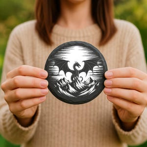 May include: A round, black slate coaster featuring a detailed white illustration of a dragon with outstretched wings atop a mountain range. The dragon design is set against a backdrop of stylized white lines, creating a dramatic contrast. The coaster is held in front of a blurred background.