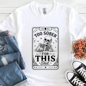 K&ouml;nnte beinhalten: Wei&szlig;es T-Shirt mit einer schwarz-wei&szlig;en Grafik eines Skeletts, das ein Getr&auml;nk h&auml;lt, mit dem Text "TOO SOBER FOR THIS Shit". Das Shirt wird mit blauen Jeans und schwarz-wei&szlig;en Turnschuhen gezeigt.